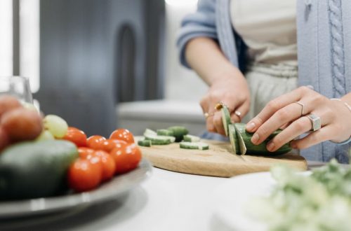Close-up of a woman slicing cucumbers on a wooden board surrounded by fresh vegetables.