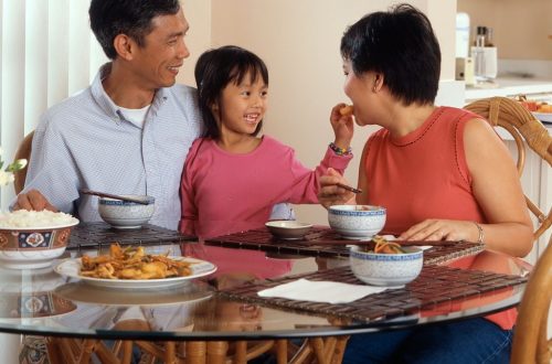 An Asian family, an adult male and female are seated around a table eating a meal with a young female standing in between the adults.